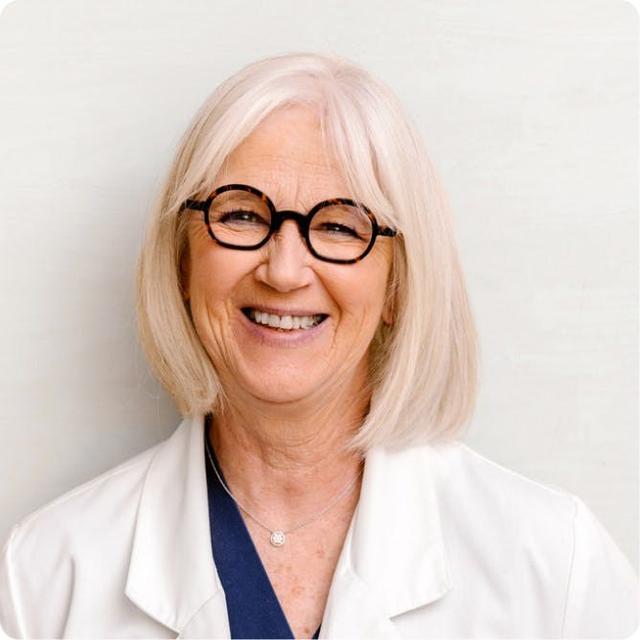 Portrait of a smiling older female healthcare provider with shoulder-length white hair, round black glasses, and a white coat.