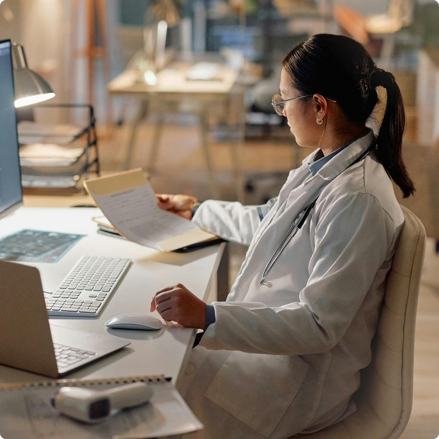 Female doctor in a white lab coat sits at a desk reviewing a patient's chart, with a stethoscope around her neck and a computer monitor in front of her.