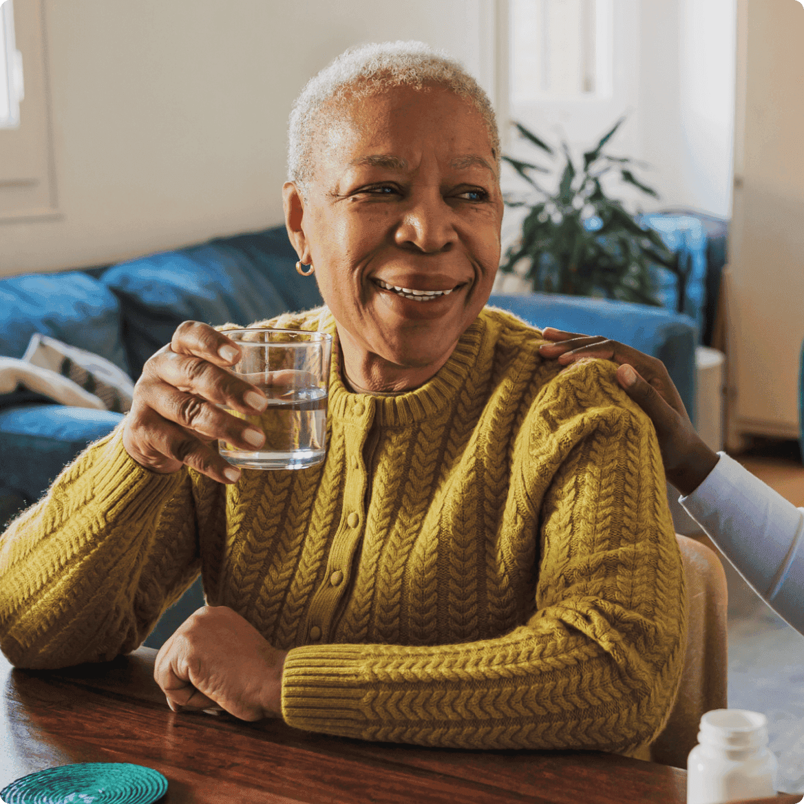 An older woman wearing a mustard-yellow knit sweater smiles while holding a glass of water at a table. A supportive hand rests on her shoulder, and a bottle of medication is visible in the foreground, suggesting a healthcare or caregiving context.