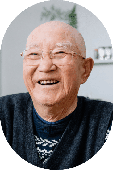 Smiling elderly man with glasses and a bald head, wearing a dark sweater with a patterned collar, seated indoors.