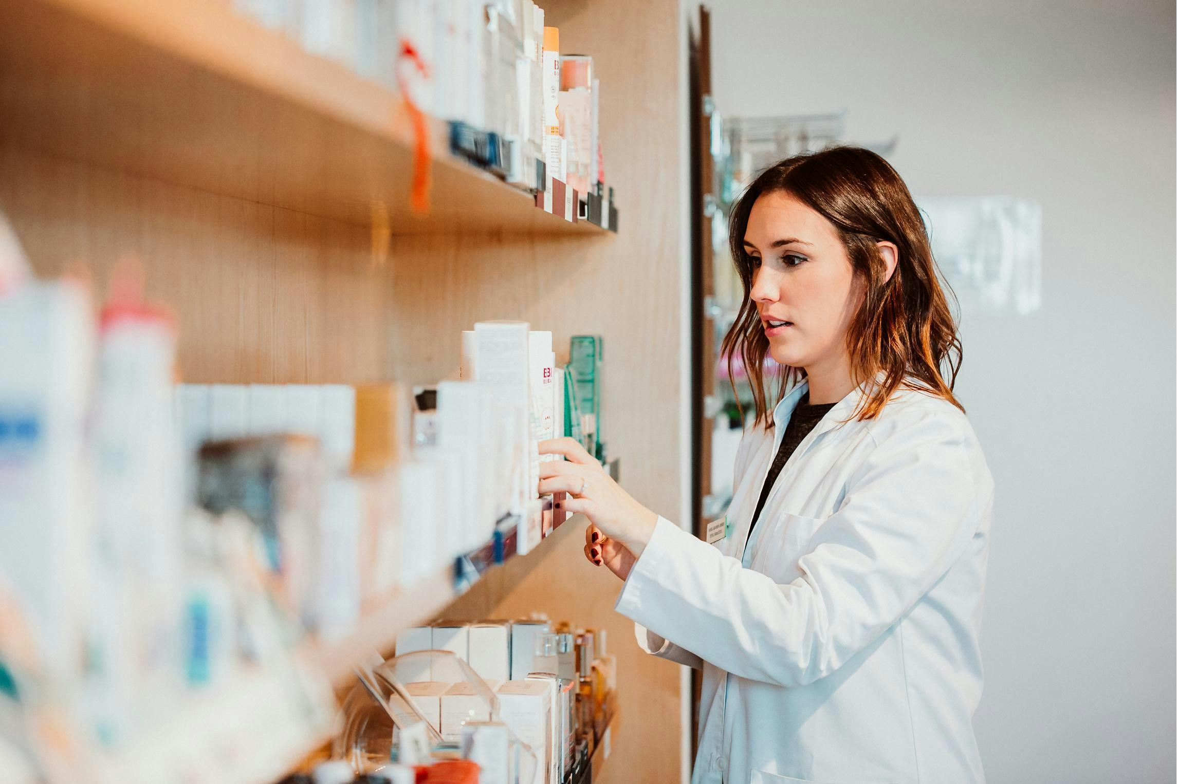 Woman in a lab coat standing in front of a counter selecting a medication for a patient.