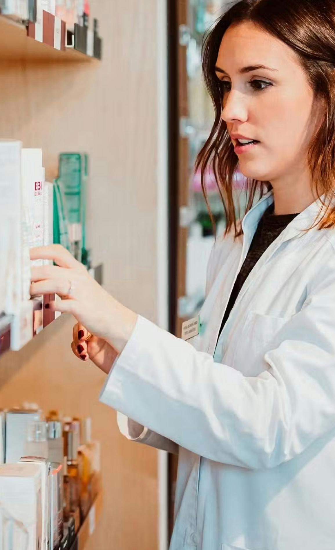 Woman in a lab coat standing in front of a counter selecting a medication for a patient.